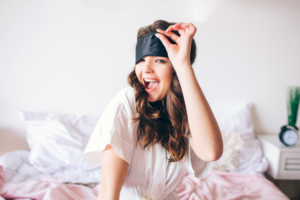 Bride to Be dressed in pink sitting in bed and lifting up her sleep eye mask.